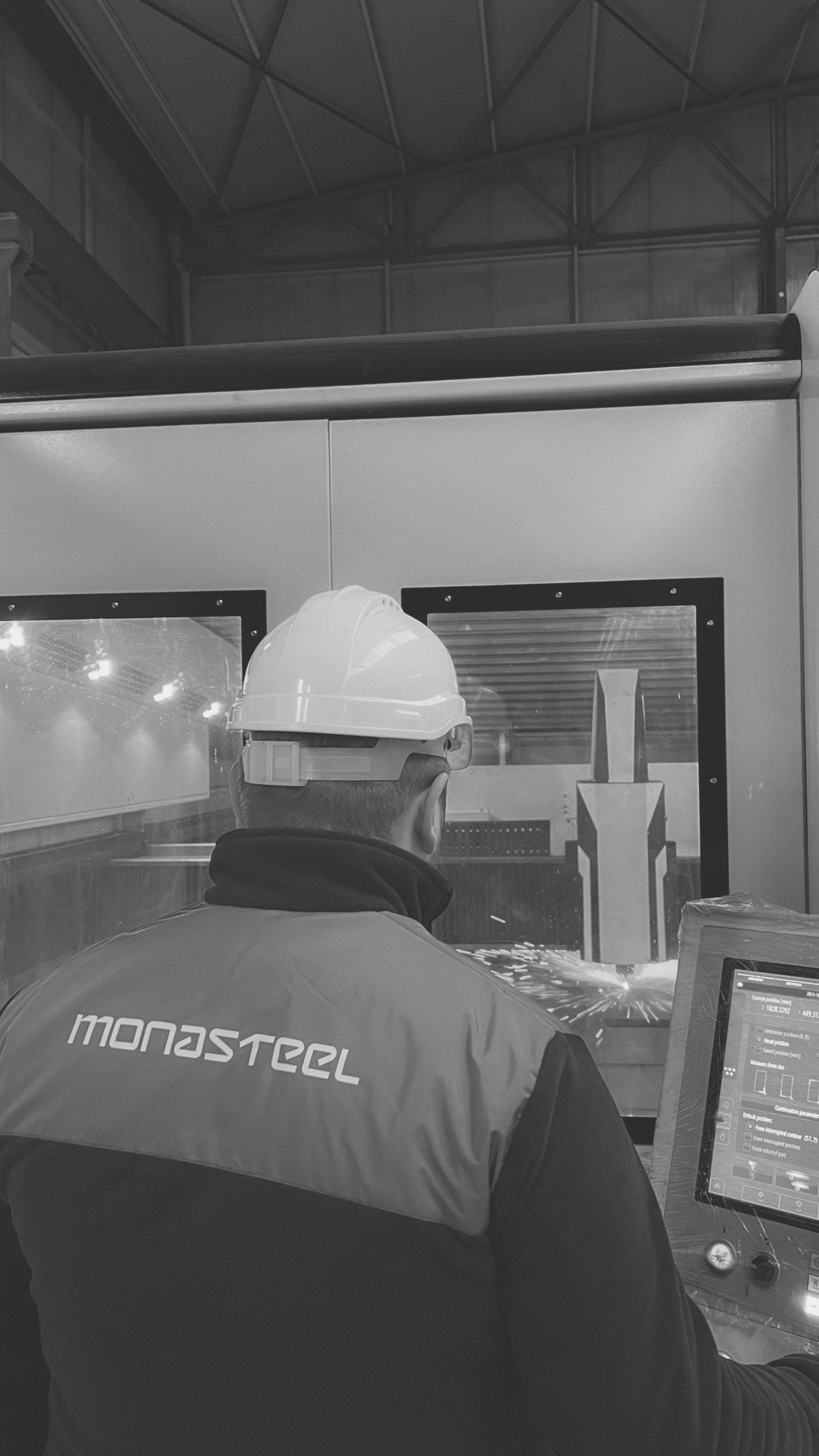 Mona Steel operator at a fiber laser under a steel truss roof, the MONASTEEL wordmark across the back of his jacket